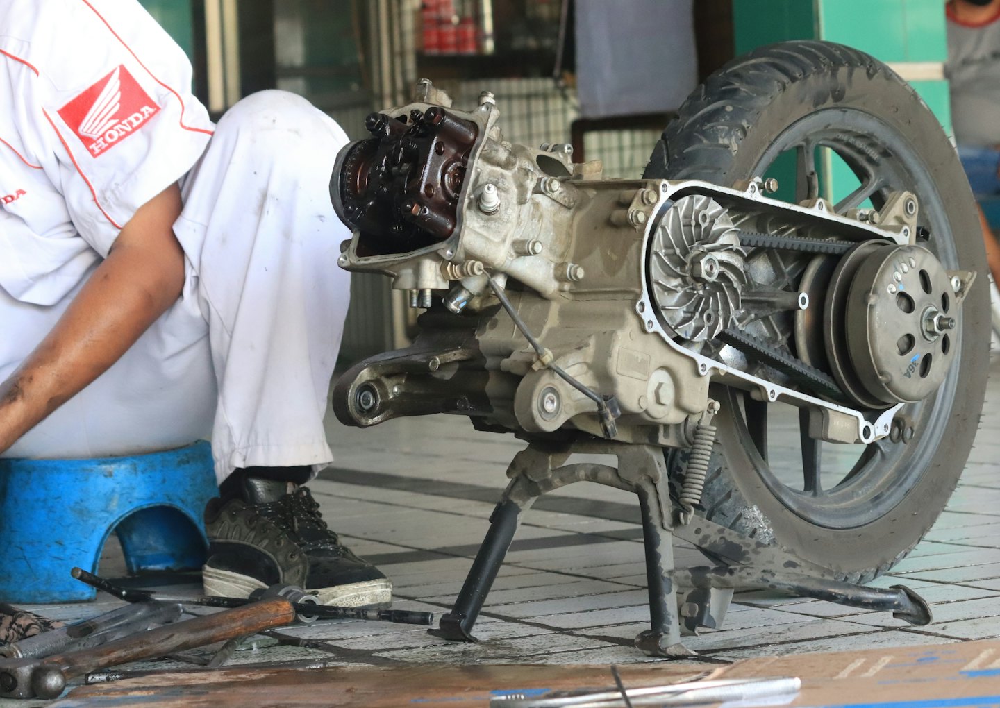 Mechanic works on scooter engine with tools on floor.